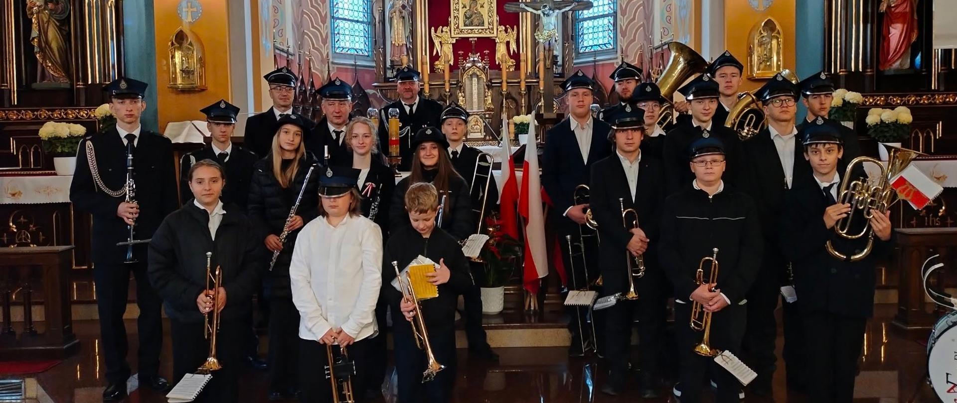 Na zdjęciu widać grupę kilkudziesięciu osób stojących w kościele, przed ołtarzem. To orkiestra dęta — członkowie mają instrumenty muzyczne, takie jak trąbki, klarnety, puzony, sakshorny i bębny. Większość osób ubrana jest w ciemne mundury z czapkami, część młodszych w białych koszulach lub ciemnych kurtkach. Za nimi znajduje się bogato zdobiony ołtarz z krzyżem, figurami świętych i licznymi złotymi elementami. W tle widoczne są kolorowe witraże i obrazy religijne, między innymi przedstawienie Jana Pawła II i Jezusa Miłosiernego. Przed grupą, na posadzce, stoi rząd zapalonych czerwonych zniczy. Zdjęcie ma uroczysty, podniosły charakter.