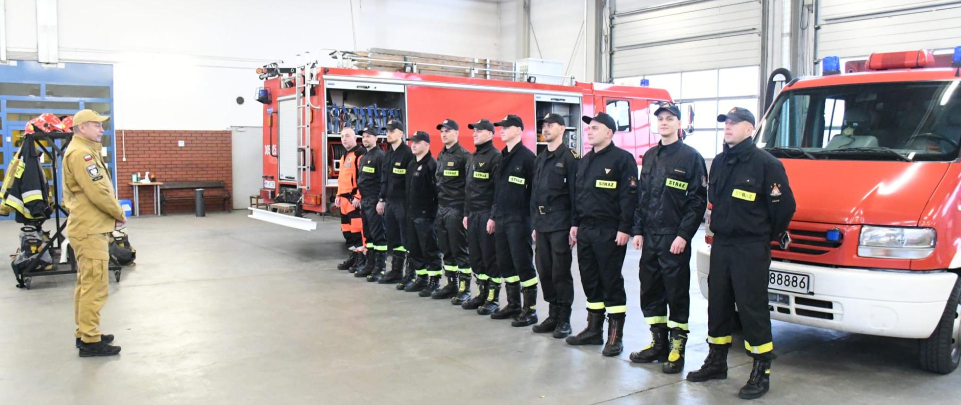 Z prawej strażacy stojący w szeregu w czarnym ubraniu koszarowym, z lewej zastępca Komendanta Głównego w żółtym ubraniu służbowym. 