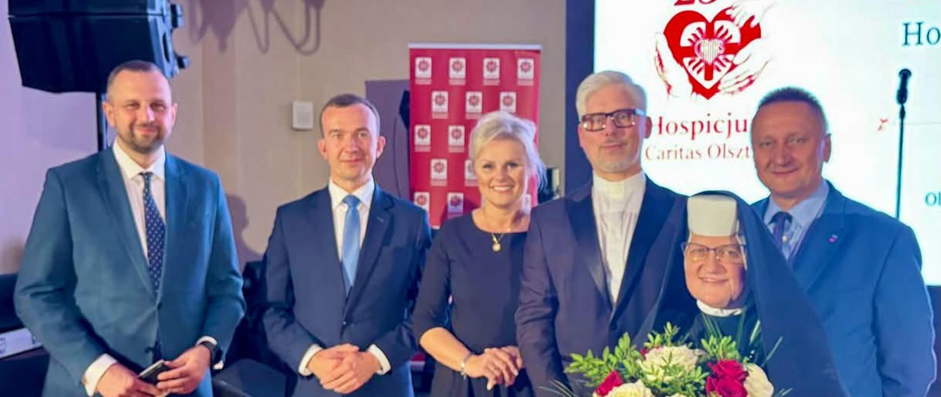 Grupa oficjeli pozuje wspólnie do zdjęcia na tle grafik przedstawiających Hospicjum Caritas w Olsztynie.