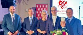 Grupa oficjeli pozuje wspólnie do zdjęcia na tle grafik przedstawiających Hospicjum Caritas w Olsztynie.