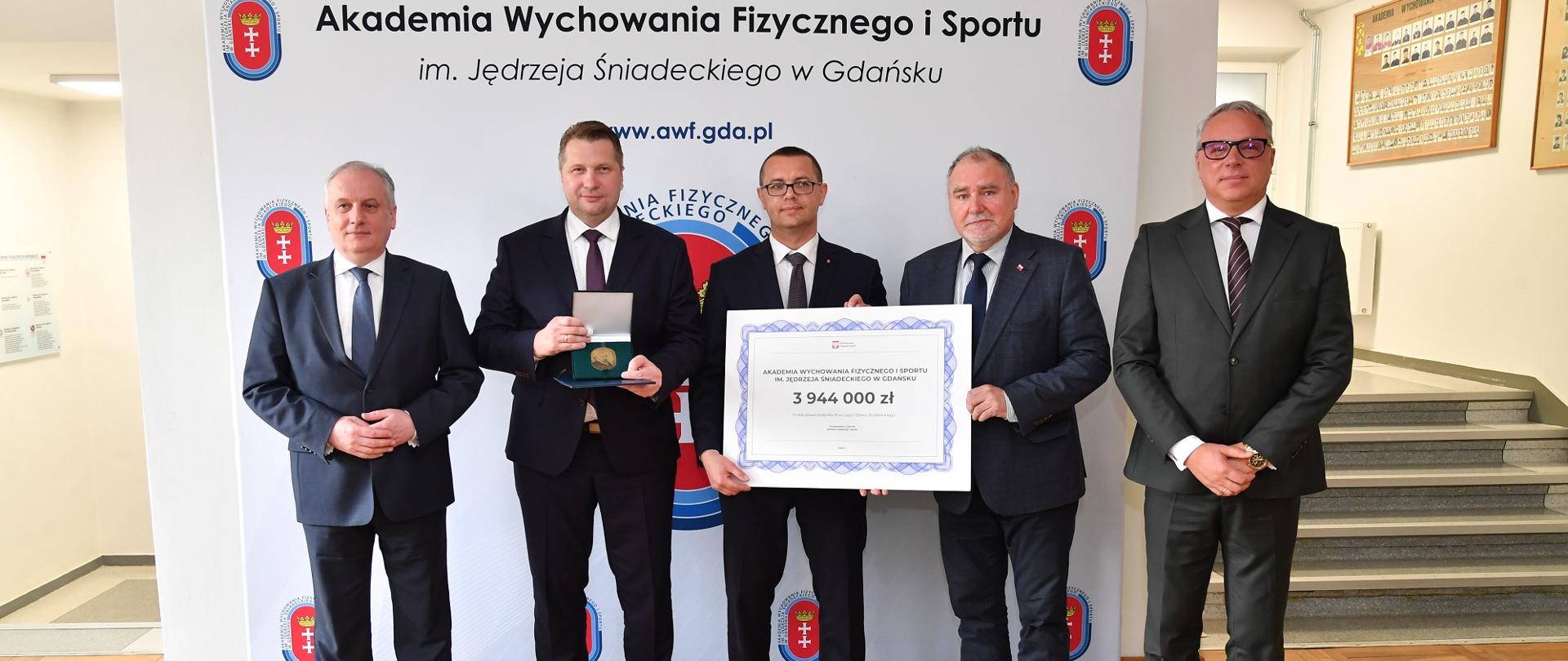 Uczestnicy wizyty stoją przy schodach, jeden trzyma symboliczny czek.