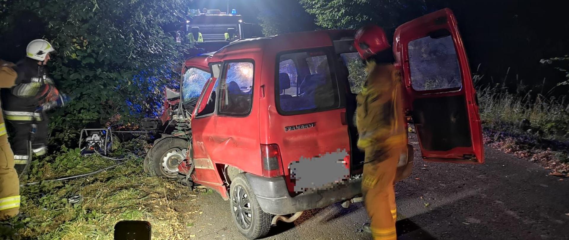 Noc, samochód strażacki oświetla teren, widoczny czerwony samochód, który ma rozbity przód.