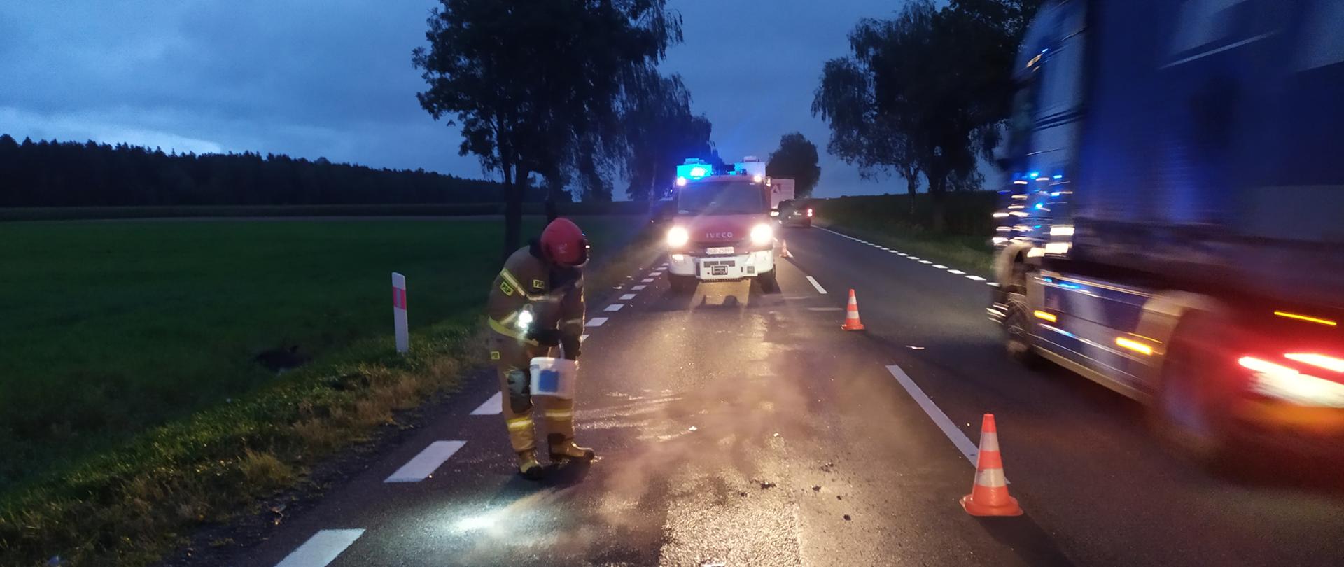 Strażak posypuje sorbentem jezdnię. W tle samochód strażacki z prawej jedzie ciężarówka. Dookoła zmrok.