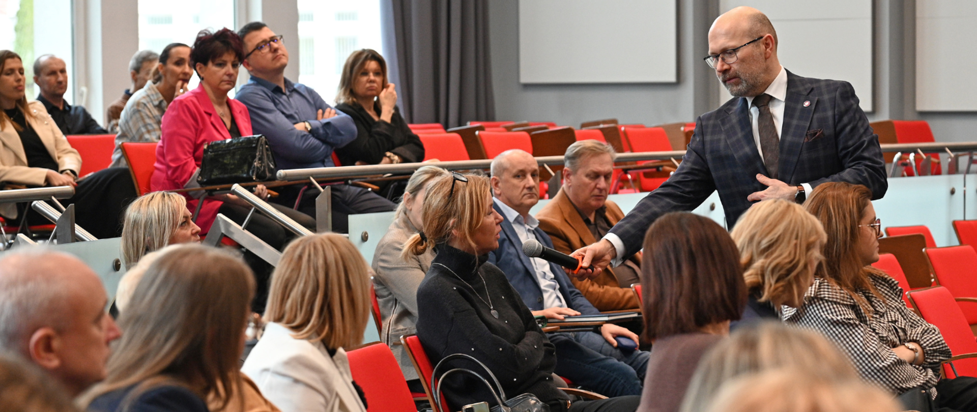 Mężczyzna w garniturze podaje mikrofon siedzącej kobiecie podczas spotkania lub konferencji w sali wykładowej. W tle widać publiczność siedzącą na czerwonych krzesłach, słuchającą dyskusji.