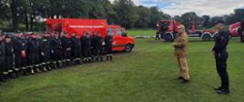 strażak w jasnym mundurze stoi przed grupą strażaków w czarnych mundurach, wszyscy stoją na polanie, w tle pojazdy pożarnicze