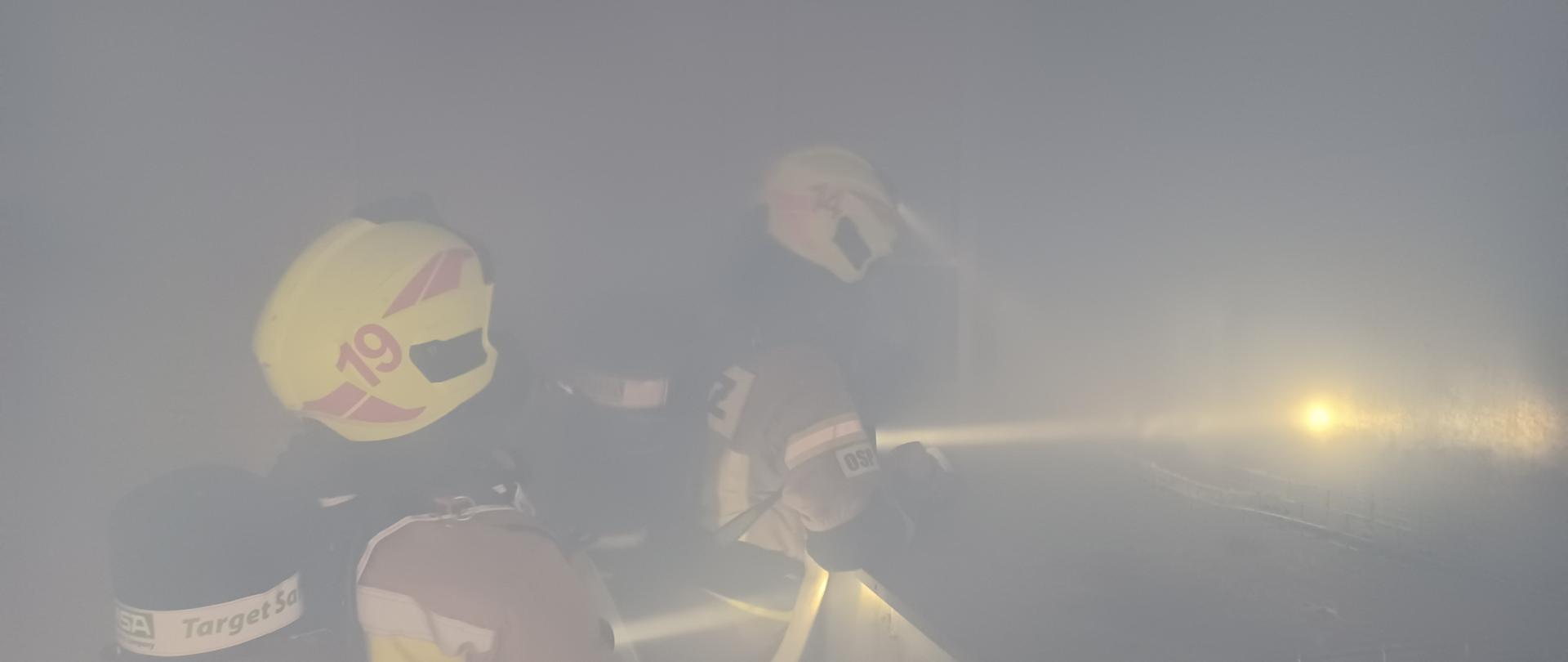 Na pierwszym planie widać strażaków w sprzęcie ochrony układu oddechowego poruszających się z linią wężową wewnątrz obiektu.