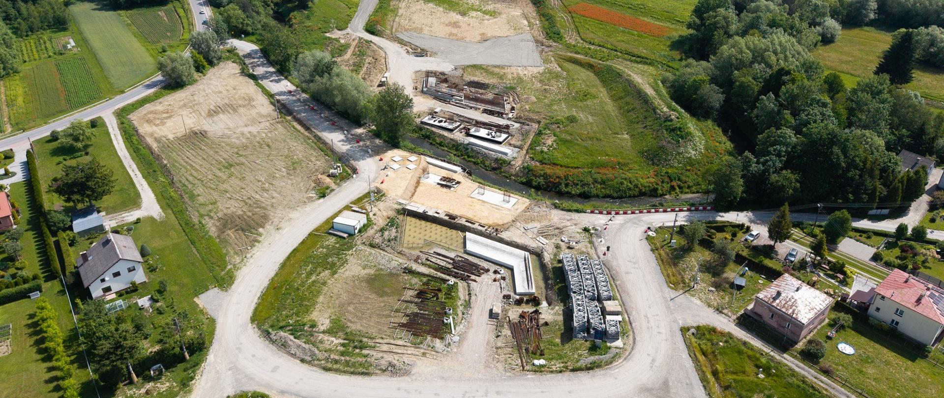 Lotnicze zdjęcie przedstawiające teren budowy drogi w otoczeniu pól uprawnych, zadrzewień i rozproszonej zabudowy jednorodzinnej. W centralnej części widoczny jest przygotowany pas terenu pod nową trasę oraz plac budowy z fundamentami i składowanymi elementami konstrukcyjnymi. Przez środek obszaru przepływa niewielki ciek wodny, nad którym powstaje obiekt mostowy. Całość otaczają zielone łąki, drzewa i lokalne drogi.
