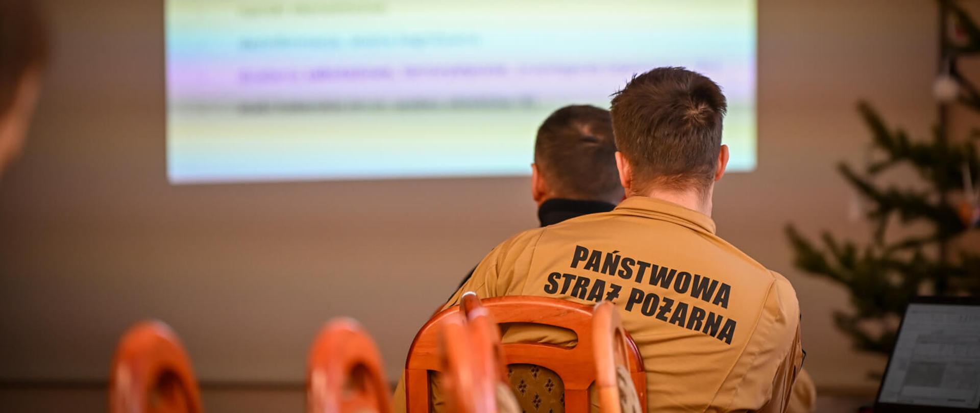 Widok zza pleców strażaka w piaskowym mundurze z napisem „PAŃSTWOWA STRAŻ POŻARNA” na plecach. Mężczyzna siedzi w sali konferencyjnej i patrzy na wyświetlaną na ekranie prezentację.