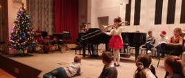 Uczennica grająca na puzonie oraz nauczyciel akompaniujący na fortepianie, w tle dzieci siedzące na scenie i ozdobiona świąteczna choinka