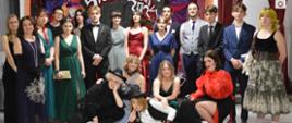 Zdjęcie grupowe, pozowane wykonane w szkolnej auli. Uczniowie w eleganckich ubraniach. Niektóre stroje posiadają elementy nawiązujące do baśni "Alicja w Krainie Czarów". Większość młodzieży stoi. Kilku uczniów jest w przysiadzie. Jeden chłopiec leży na podłodze. Jest też nauczycielka Ewa Wrona. W tle dekoracja.
