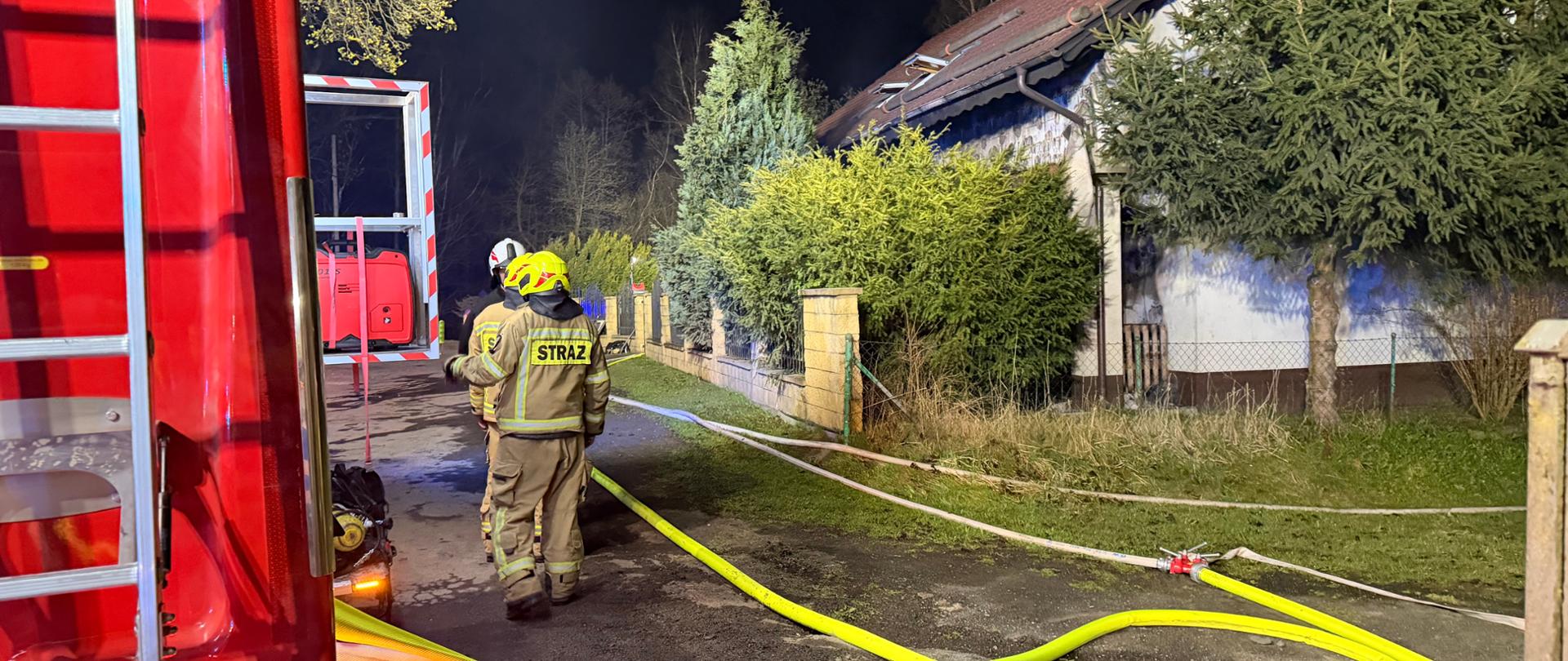 wozy strażackie, dwóch strażaków, rozwinięte węże strażackie, nic w tle dom jednorodzinny po prawej stronie