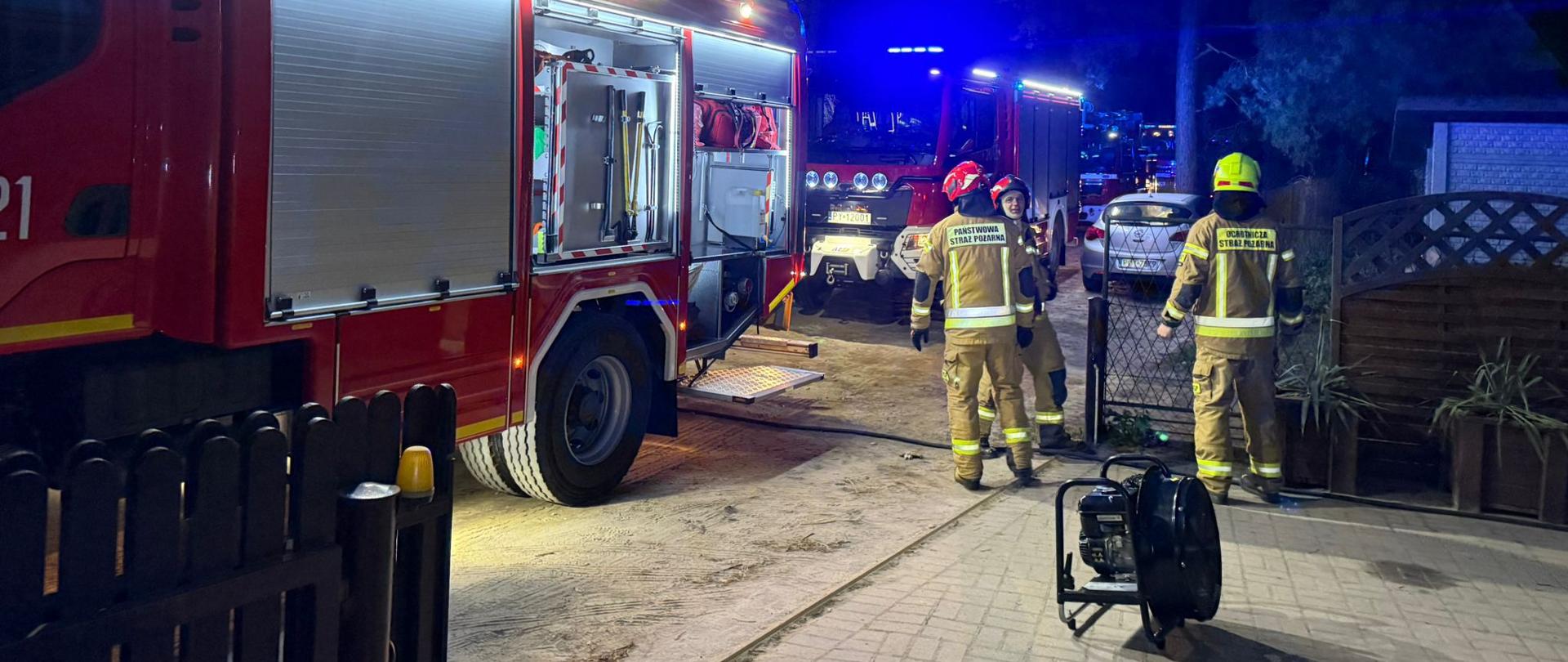 Strażacy w ubraniach specjalnych stoją w pobliżu wozów strażackich. Przy nich znajduje się sprzęt pożarniczy. W tle las i zabudowania.