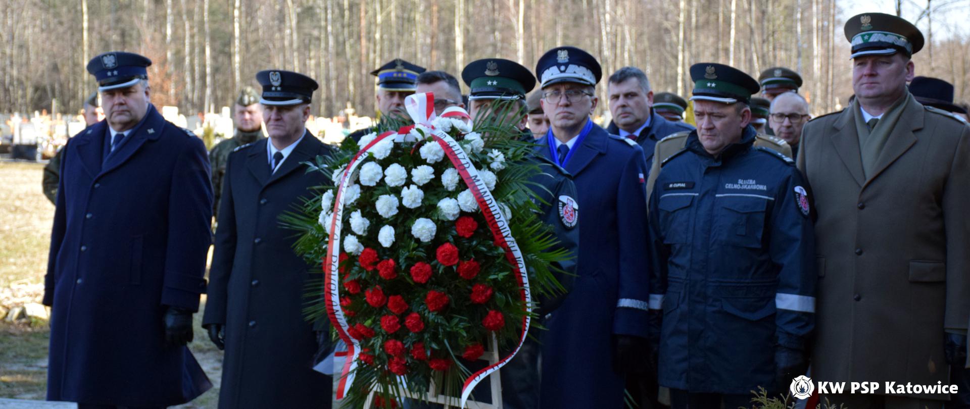 Na cmentarzu murckowskim w Katowicach, w szeregu stoją w mundurach szefowie służb mundurowych. Przed nimi na stojaku wieniec z biało-czerwonych kwiatów. 