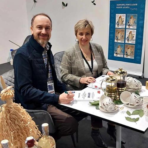 Kobieta i mężczyzna siedzą przy stoliku, na którym stoją dzbanki z herbatą i filiżanki ze spodeczkami, i podpisują certyfikaty