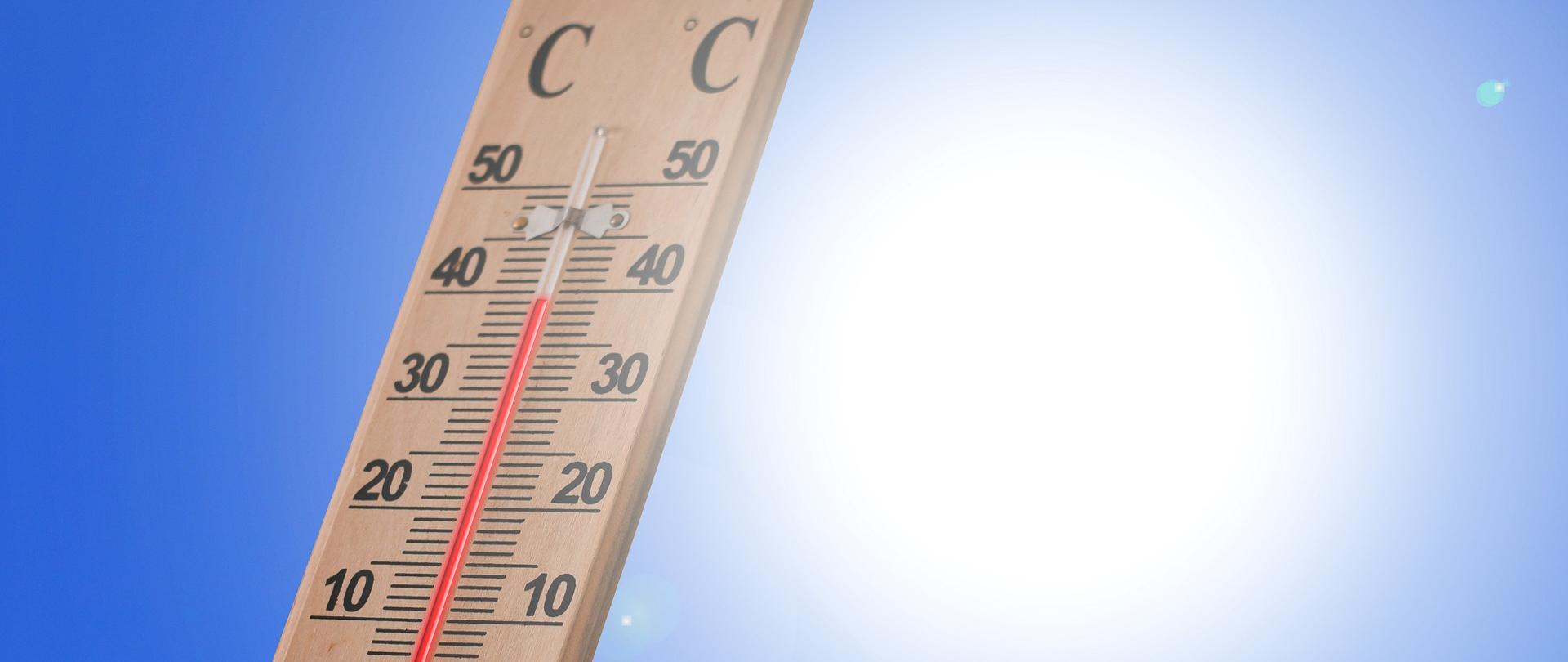 Na zdjęciu znajduje się termometr ścienny pokazujący temperaturę 40 stopni Celsjusza. W tle widać pogodne, słoneczne niebo. 