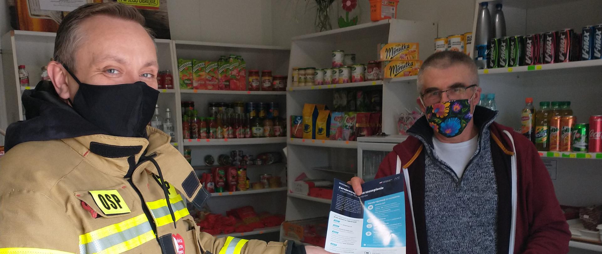 Zdjęcie przedstawia strażaka który przekazuje ulotkę na temat szczepień pracownikowi sklepu. Obaj panowie są w maseczkach.