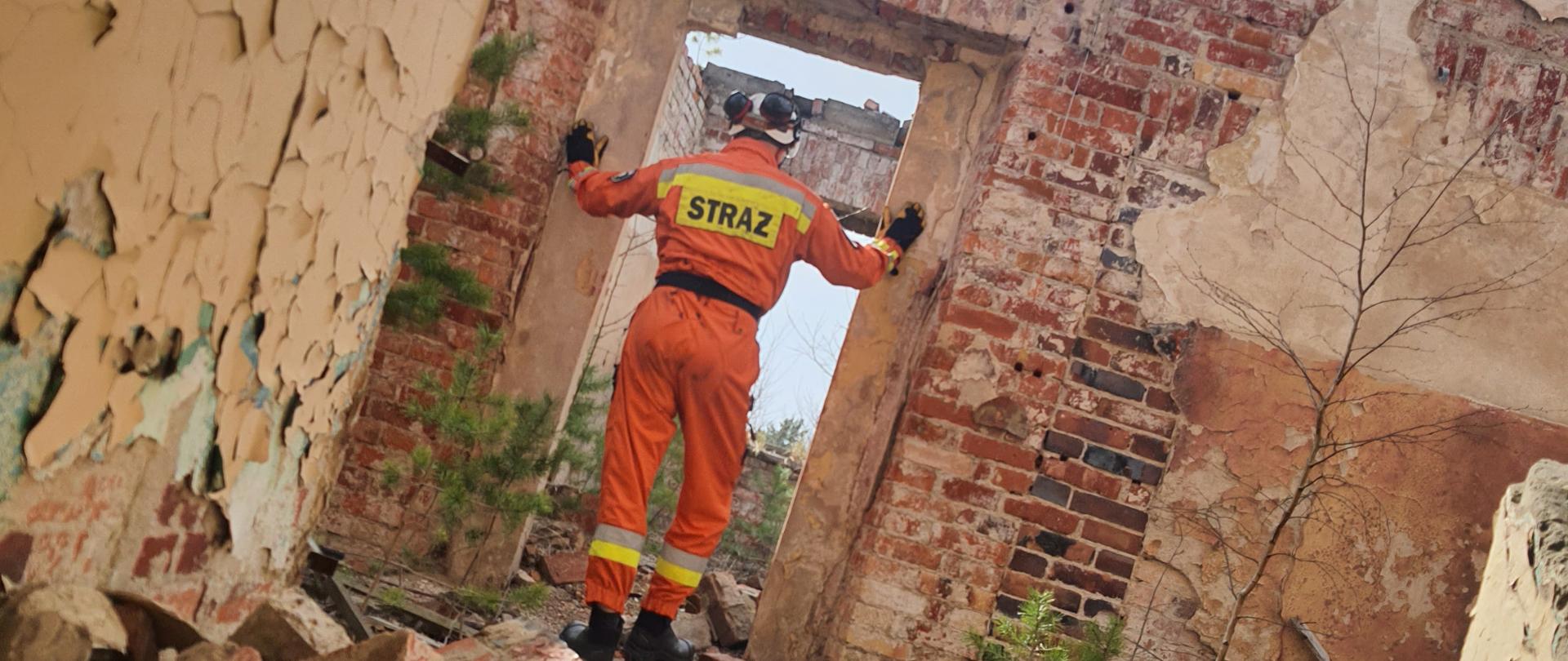 Strażak w pomarańczowym kombinezonie stojący w drzwiach częściowo zawalonego budynku