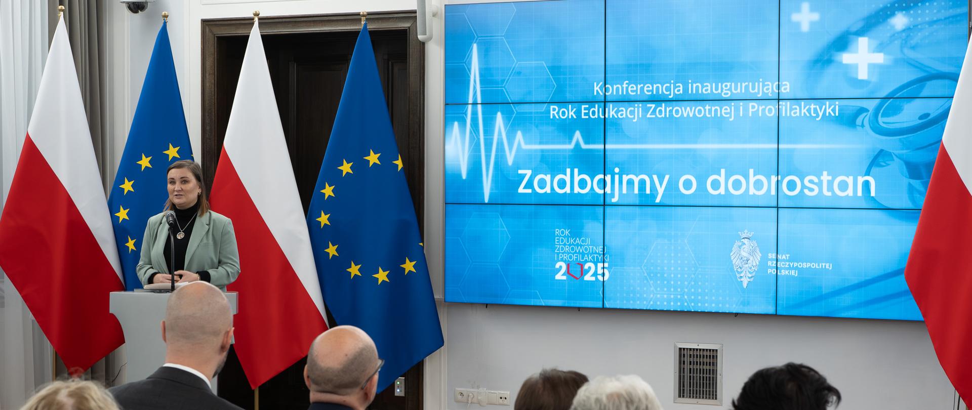 Wiceminister Paulina Piechna-Więckiewicz stoi przy mównicy i przemawia do mikrofonu. W tle znajdują się flagi Polski i UE oraz ekran, na którym wyświetlona jest nazwa konferencji "Zadbajmy o dobrostan". 