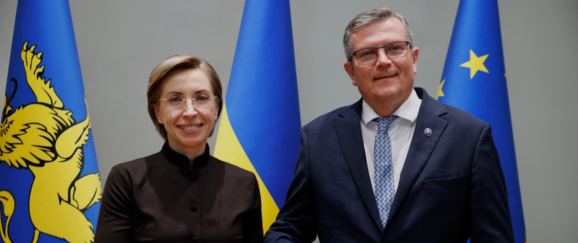 Na zdjęciu są dwie osoby - kobieta w brązowym stroju i mężczyzna w granatowym garniturze. Stoją obok siebie i patrzą w kierunku kamery. Za nimi są trzy flagi: Lwowa, Ukrainy i UE