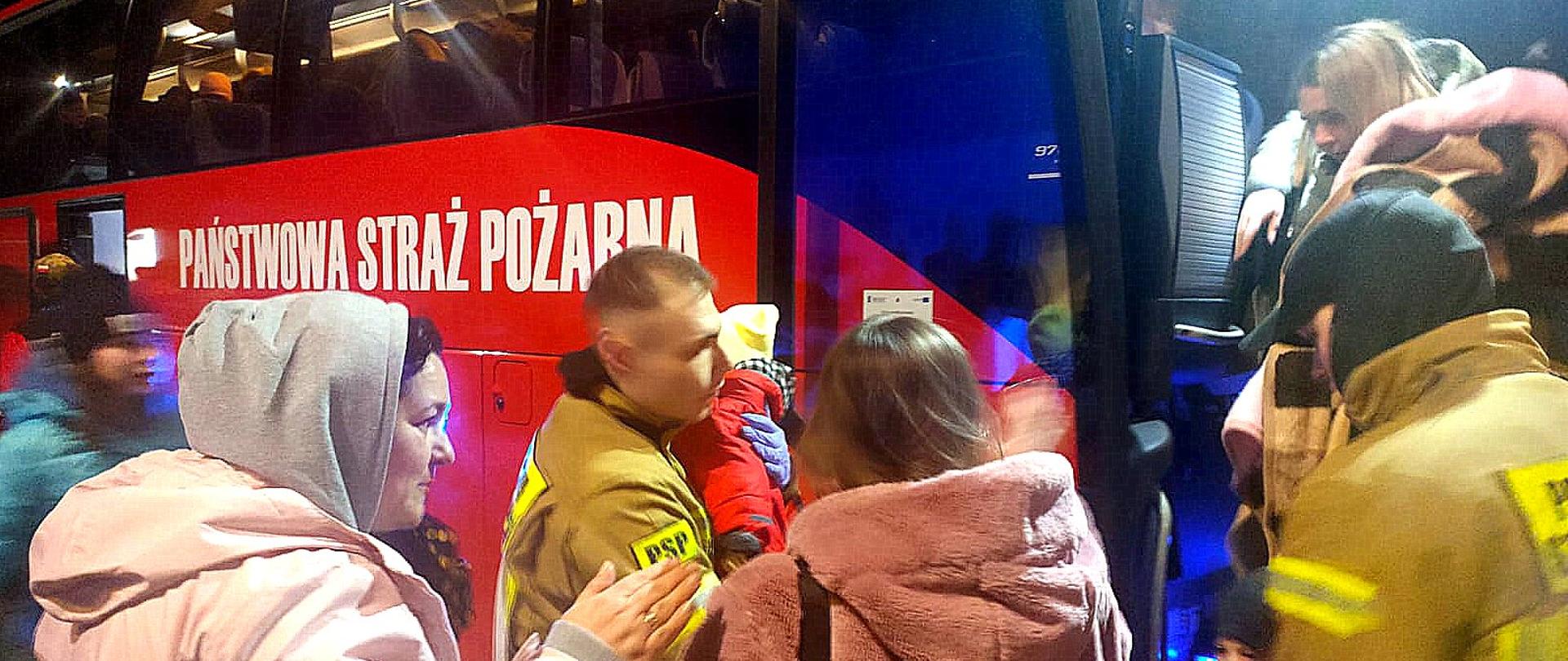 Zdjęcie zrobione na zewnątrz w nocy. Strażacy pomagają wejść do strażackiego autokaru grupie osób. Strażak trzyma na rękach małe dziecko.
