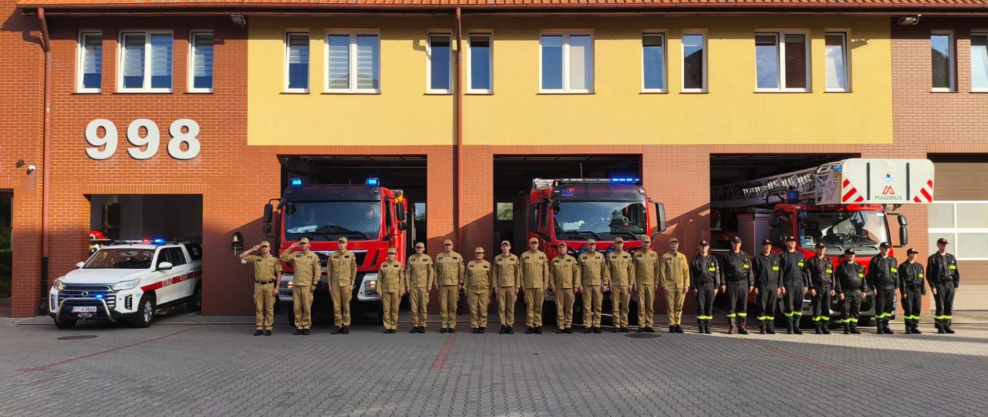 Strażacy w mundurach służbowych i bojowych stoją w szeregu przed jednostką PSP, oddając hołd poległym kolegom.