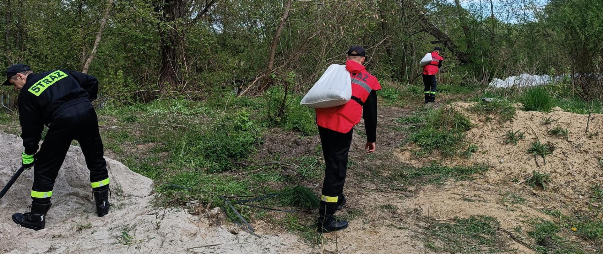 Zdjęcie przedstawia strażaków niosących worki z piaskiem w celu podwyższenia wału. Po lewo strażak napełnia kolejne worki piaskiem. W tle tereny zielone.