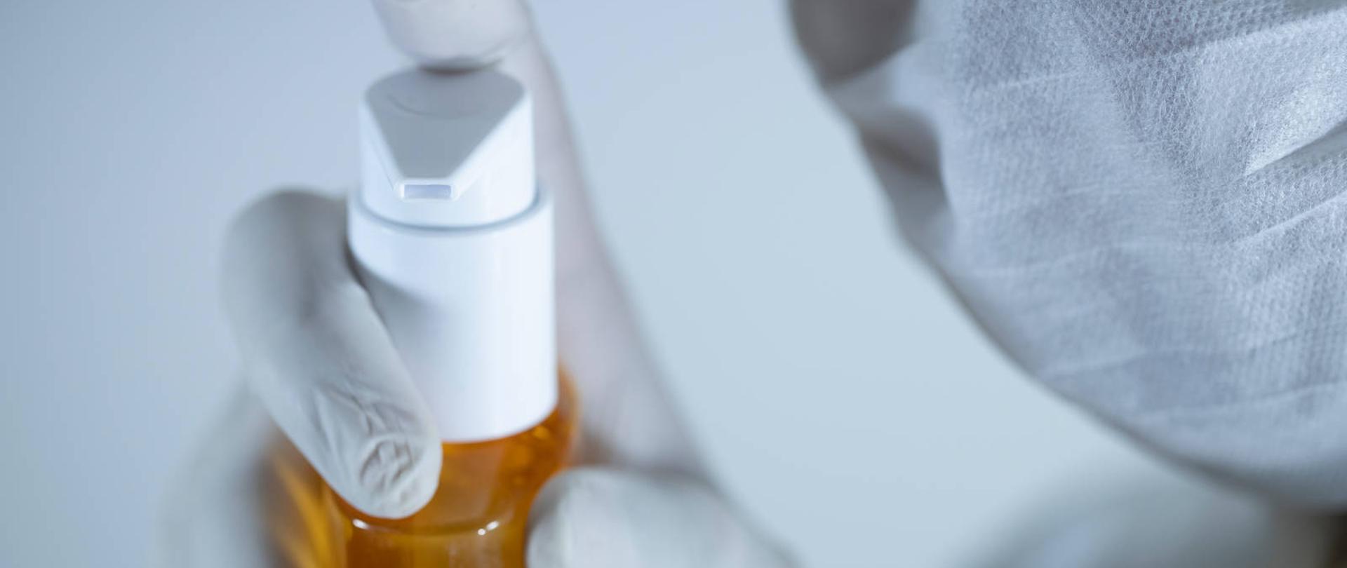 Fragment twarzy w masce i dłoń w rękawiczce na opakowaniu z płynem dezynfekującym.