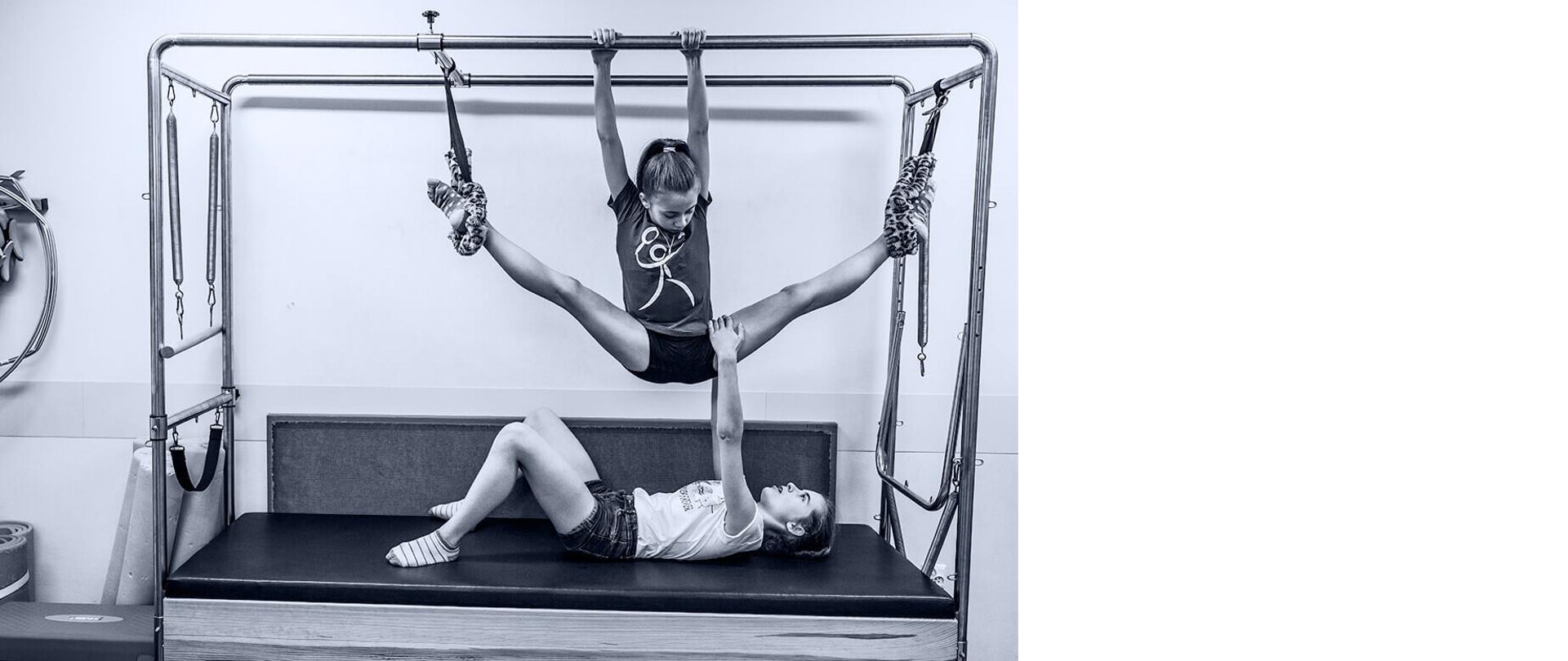 Dwie dziewczynki na łóżku do pilatesu, jedna trzymająca się drążka u góry wisząca powietrzu a druga leżąca pod nią na łóżku trzymająca za nogę wiszącą koleżankę 