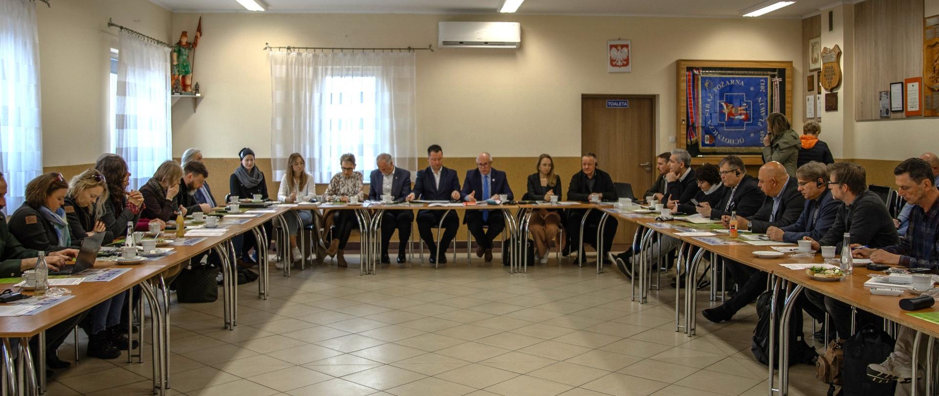 Grupa kilkunastu osób, kobiet i mężczyzn siedzi za stołem w dużej sali.