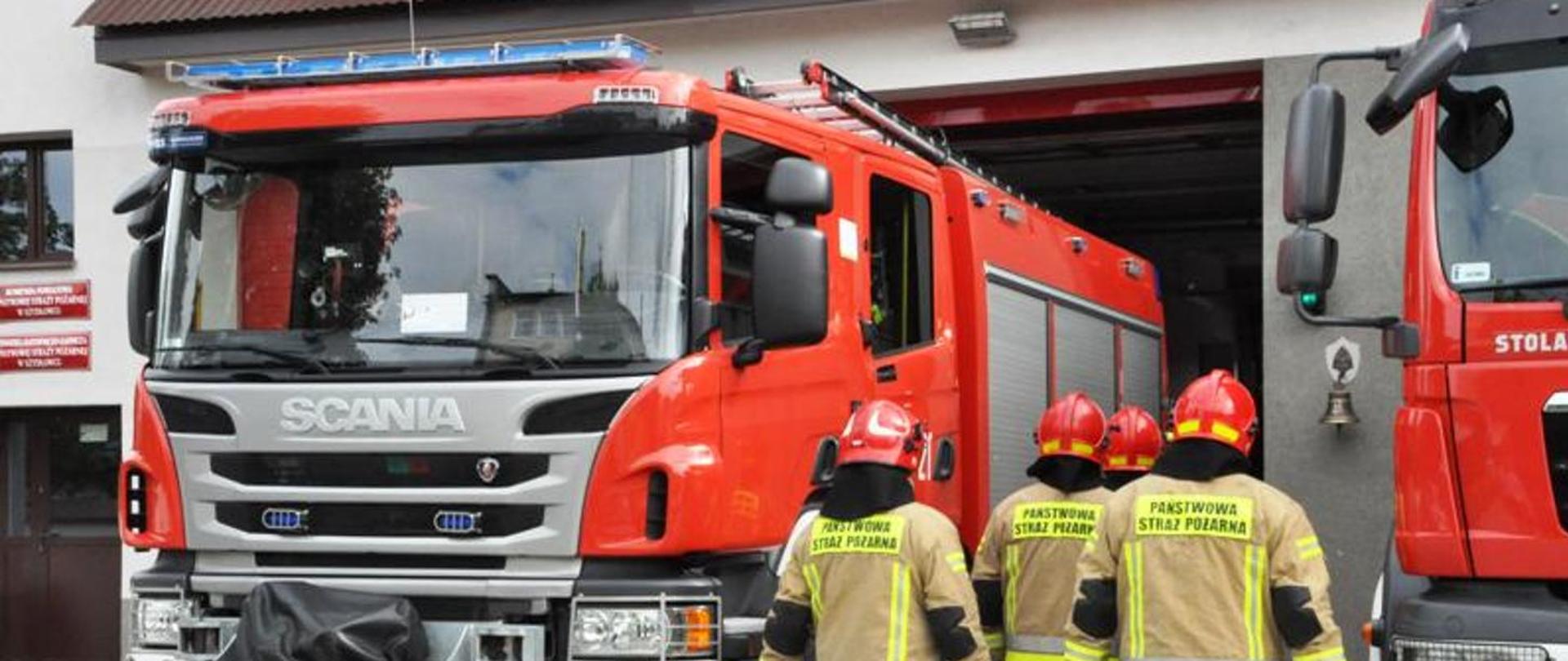 Fotografia przedstawiająca średni samochód ratowniczo-gaśniczy na tle Jednostki Ratowniczo-Gaśniczej oraz strażaków Państwowej Straży Pożarnej ubranych w ubrania specjalne .
