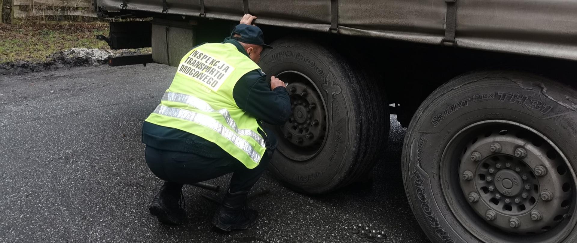 Umundurowany inspektor świętokrzyskiej Inspekcji Transportu Drogowego nachyla się przy kołach naczepy ciężarowej i kontroluje stan techniczny elementów układu hamulcowego.