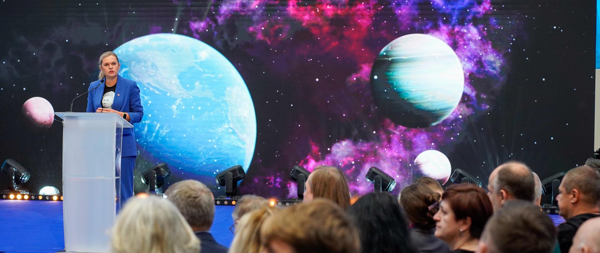 Kobieta w niebieskim garniturze przemawia z mównicy, stojąc na scenie z niebieską wykładziną. W tle znajduje się duży ekran z dynamiczną, kosmiczną grafiką – mgławicą i planetami. Publiczność siedzi w cieniu i obserwuje.