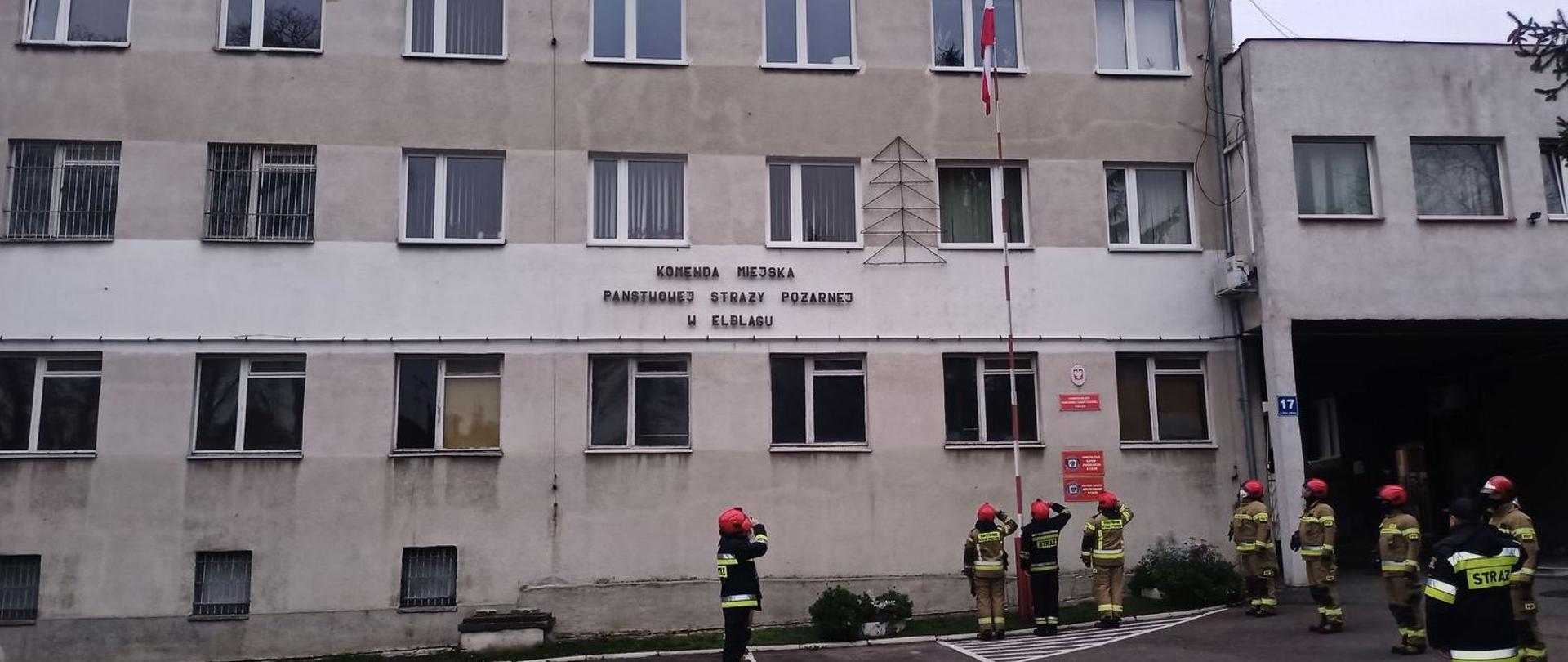 Strażacy w ubraniach specjalnych i hełmach podczas podnoszenia flagi państwowej na maszt przed siedzibą Komendy Miejskiej Państwowej Straży Pożarnej w Elblągu. Na budynku widoczny napis STRAŻ POŻARNA 998.