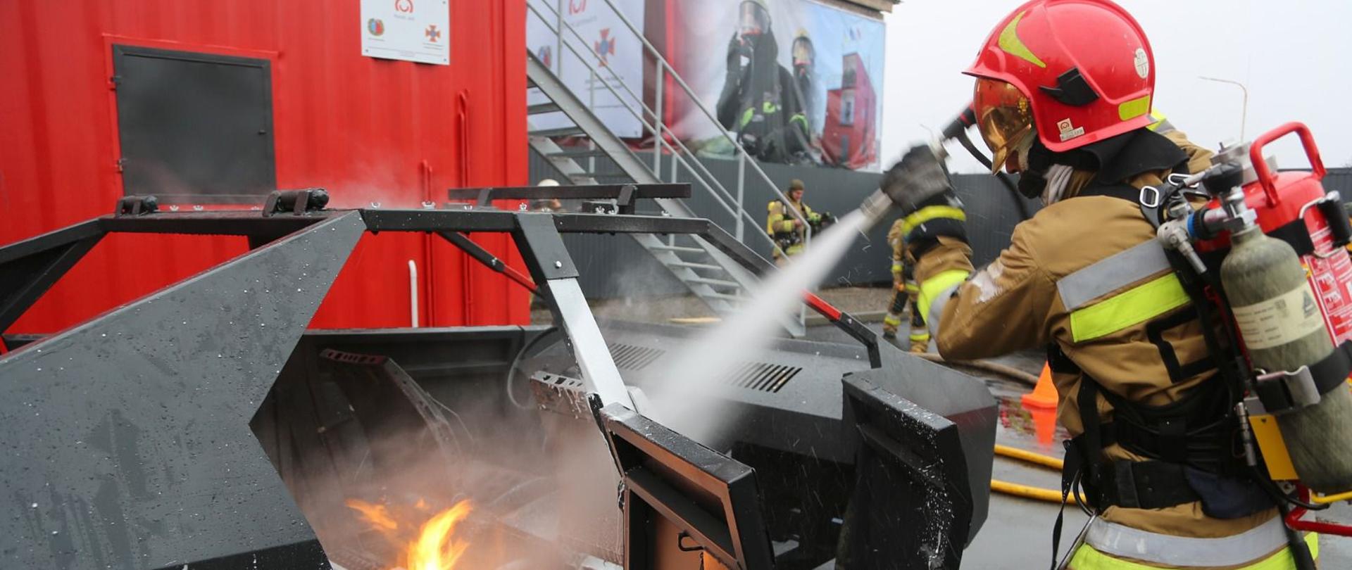 Strażak gasi płonący samochód gaśnicą, w tle logo Polska pomoc