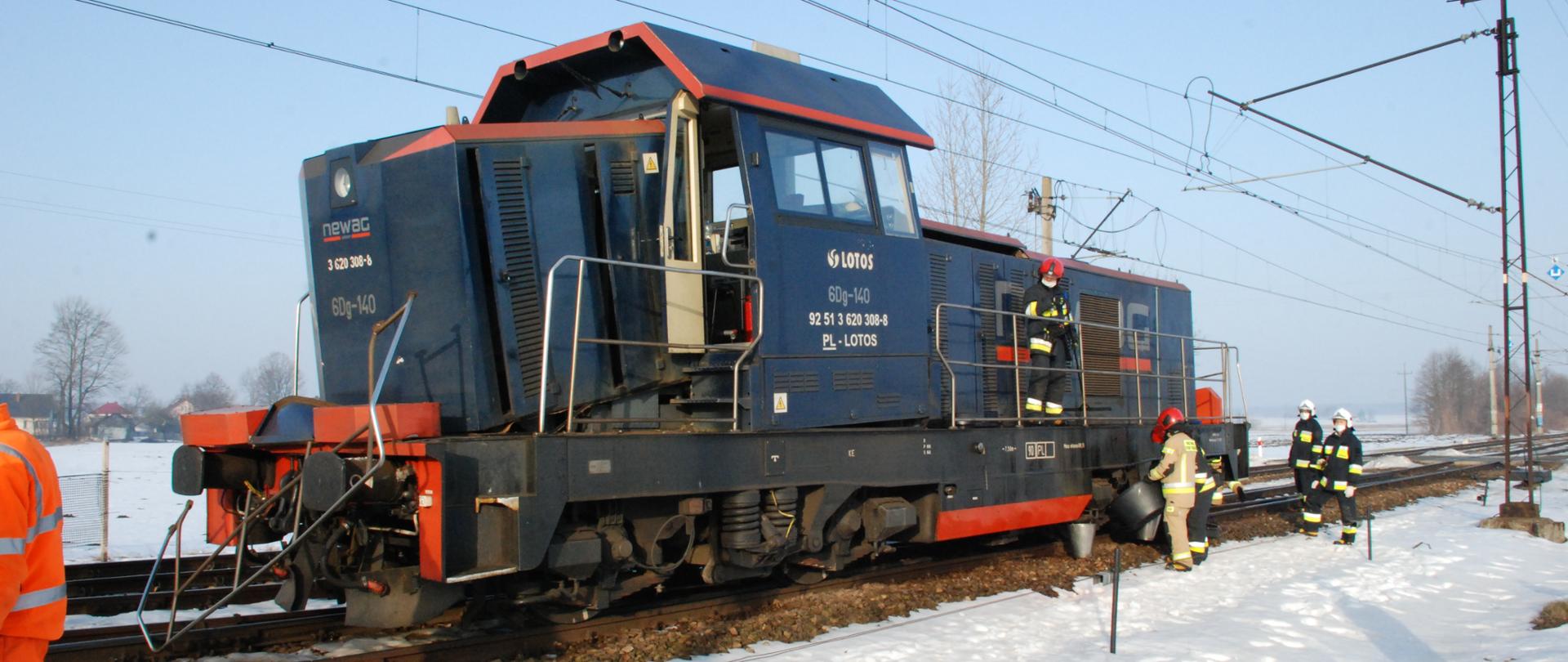 Na zdjęciu widzimy lokomotywę stojąca na torach, która została uszkodzona w skutek zderzenia z pociągiem pasażerskim. Strażacy, co jest widoczne po prawej stronie zdjęcia, zbierają i zabezpieczają wyciek oleju, który wydobywa się z lokomotywy.
