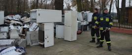 Strażacy dla Tosi. Plac komendy w Rawiczu. Na nim złożone są elektrośmieci w postaci m.in lodówek, monitorów komputerowych, drukarek, itp. Obok stoją dwaj strażacy oczekujący na kontener, do którego będą ładować towar. W tle płot i drzewa.