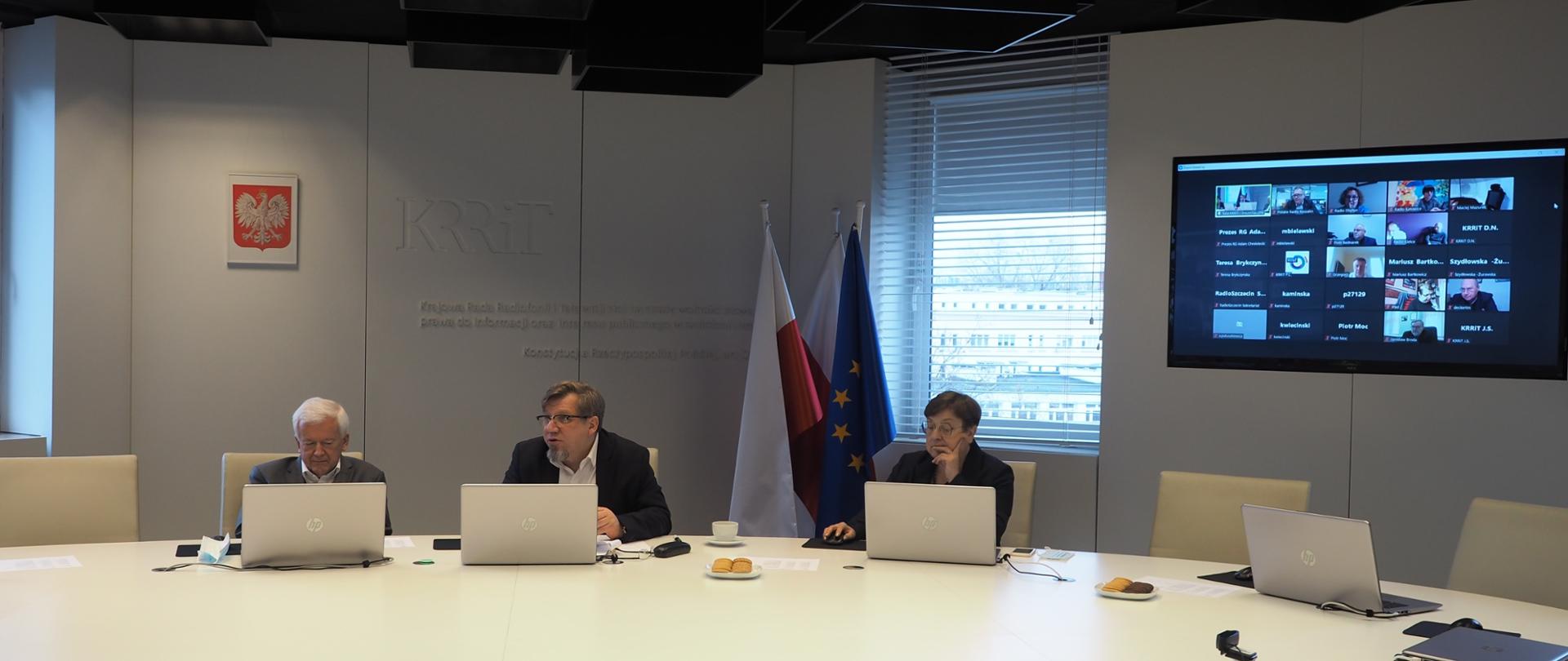 Zdjęcie ilustracyjne w poziomie. Przy owalnym białym stole siedzą Członkowie KRRiT, w tle siana z napisem "KRRiT", godło i flaga państwowa. Po prawej stronie na ścianie wisi ekran.