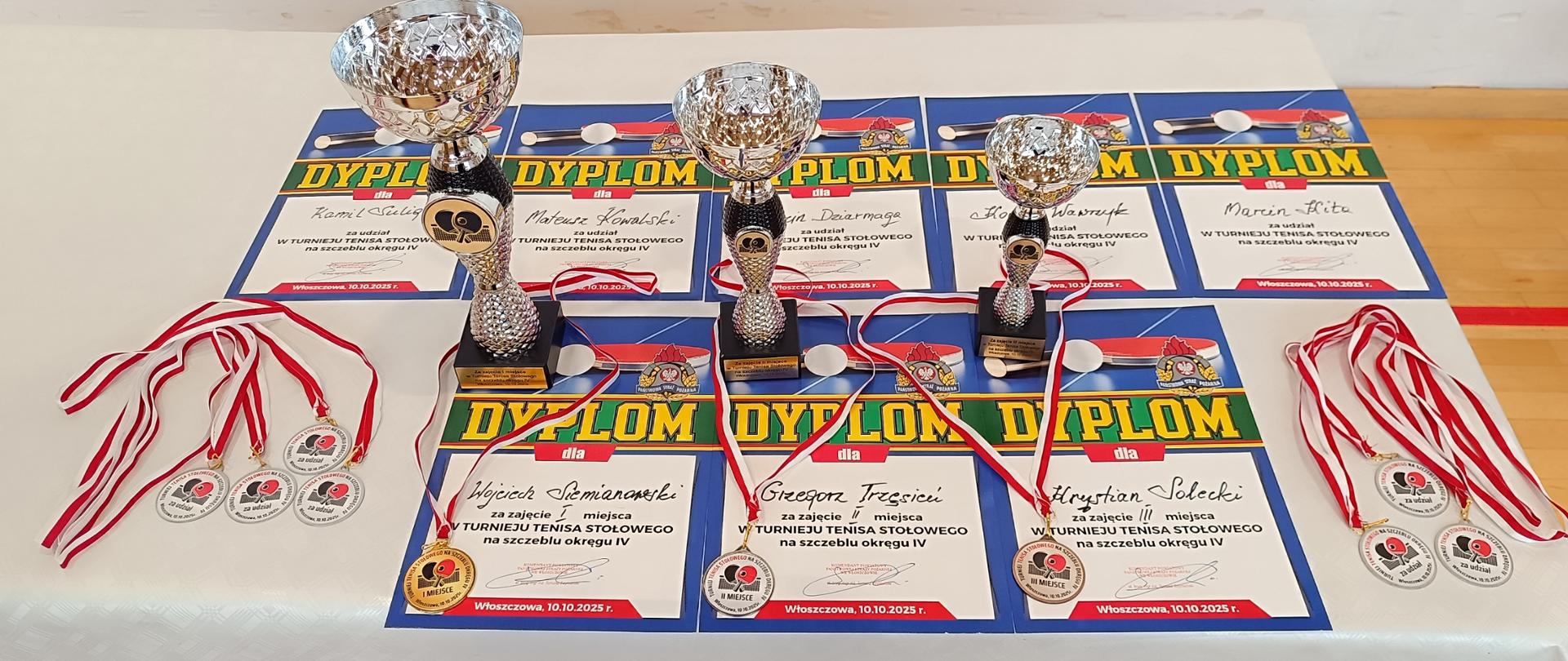 Na zdjęciu jest stół na którym znajdują się puchary, dyplomy oraz medale dla uczestników rozgrywek tenisa stołowego.