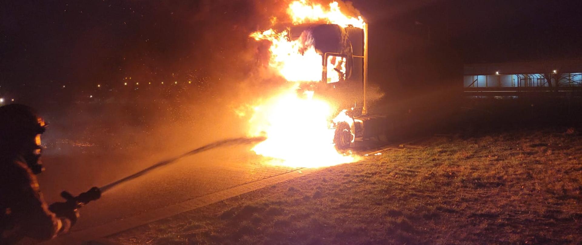 Na zdjęciu widoczny palący się samochód ciężarowy. Samochód zaparkowany na parkingu na kostce brukowej. Po lewej stronie widoczny strażak podający prąd gaśniczy na ognisko pożaru.