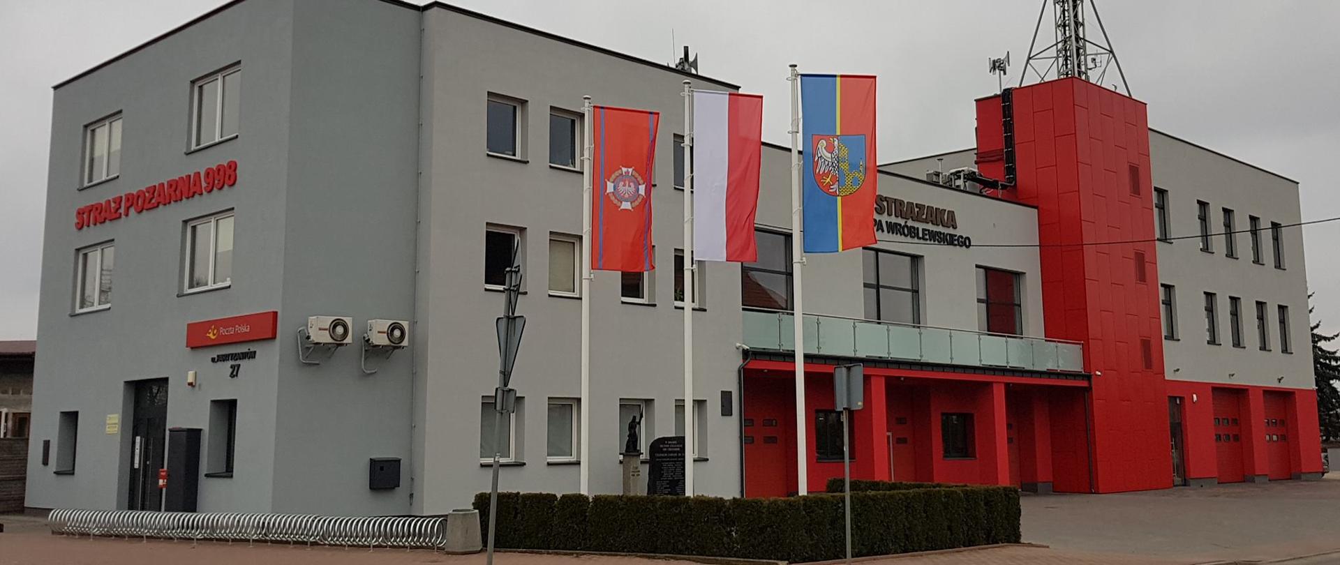 Strażnica Ochotniczej Straży Pożarnej w Chotomowie gm. Jabłonna Na pierwszym olanie 3 maszty flagowe z w tle budynek OSP. Pietrowy z szaro czerwoną elewacją. Na dole bramy garażowe na dachu maszt antenowy.