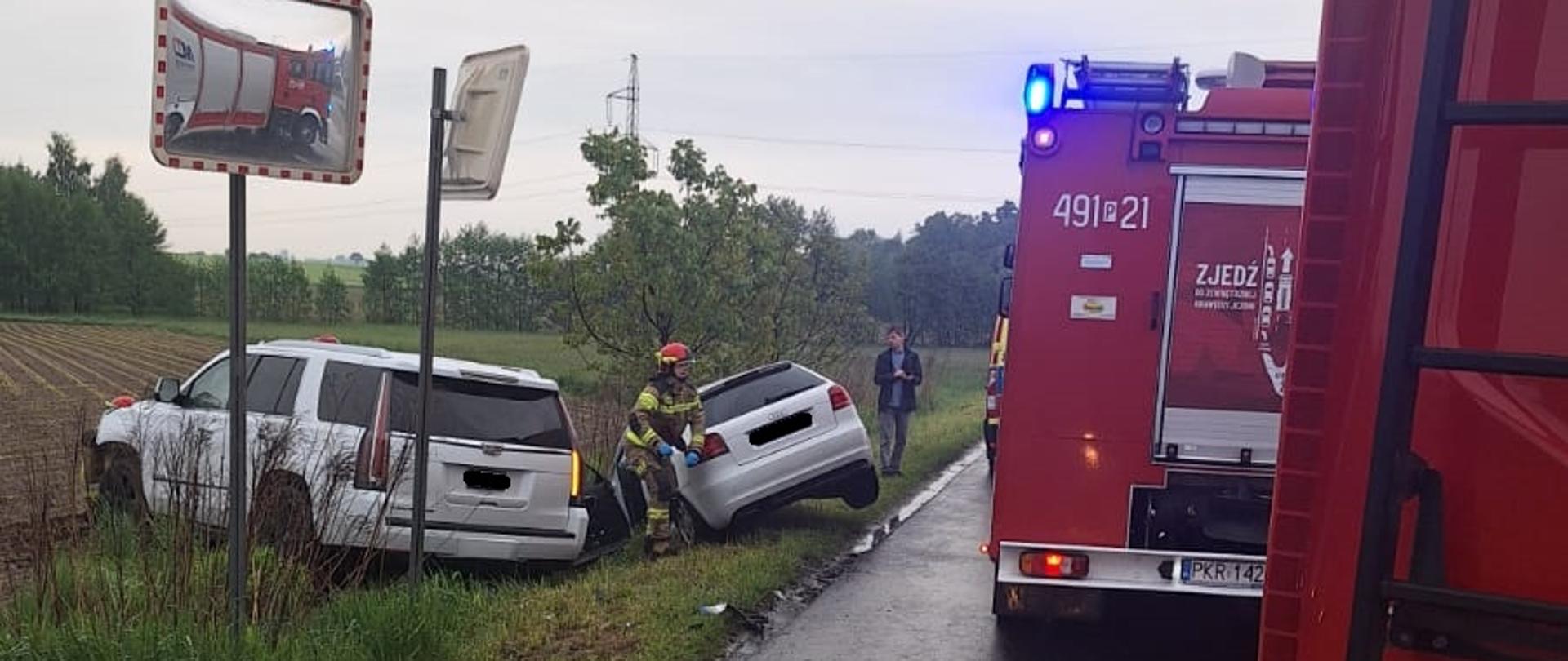Zdjęcie przedstawia dwa pojazdy osobowe koloru białego w rowie po zdarzenia drogowym. w tle samochody pozarnicze. 