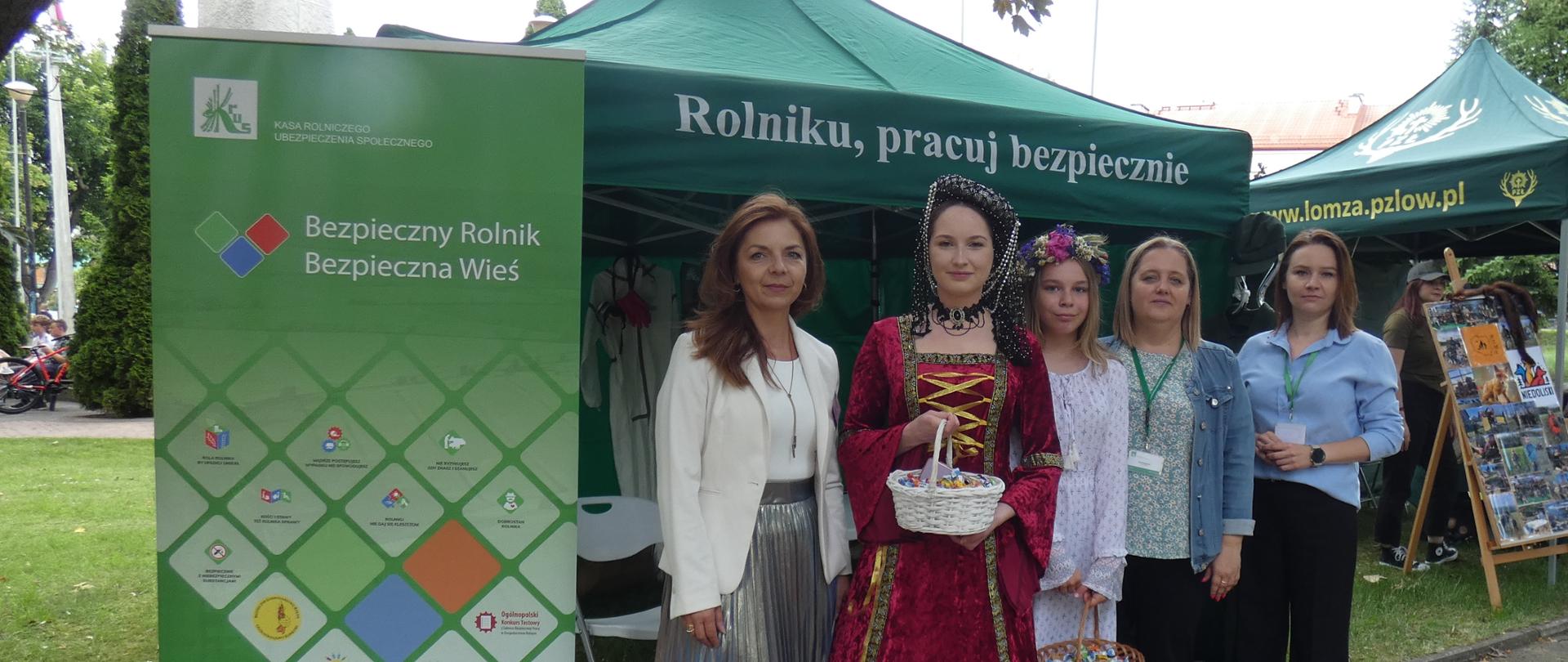 Grupa pięciu kobiet, w tym kierownik Placówki Terenowej KRUS w Łomży, stoi przed zielonym namiotem z logo KRUS i hasłem „Rolniku, pracuj bezpiecznie”. Obok widoczny jest baner promujący kampanię „Bezpieczny Rolnik, Bezpieczna Wieś”. Jedna z kobiet ubrana jest w stylizowany, historyczny strój i trzyma koszyczek ze słodyczami.