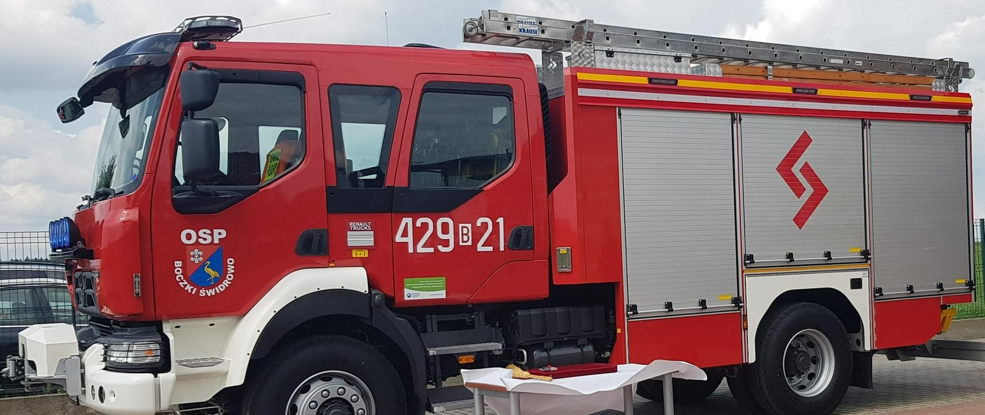 Nowy samochód gaśniczy OSP stoi na placu. Przed nim stolik z białym nakryciem.