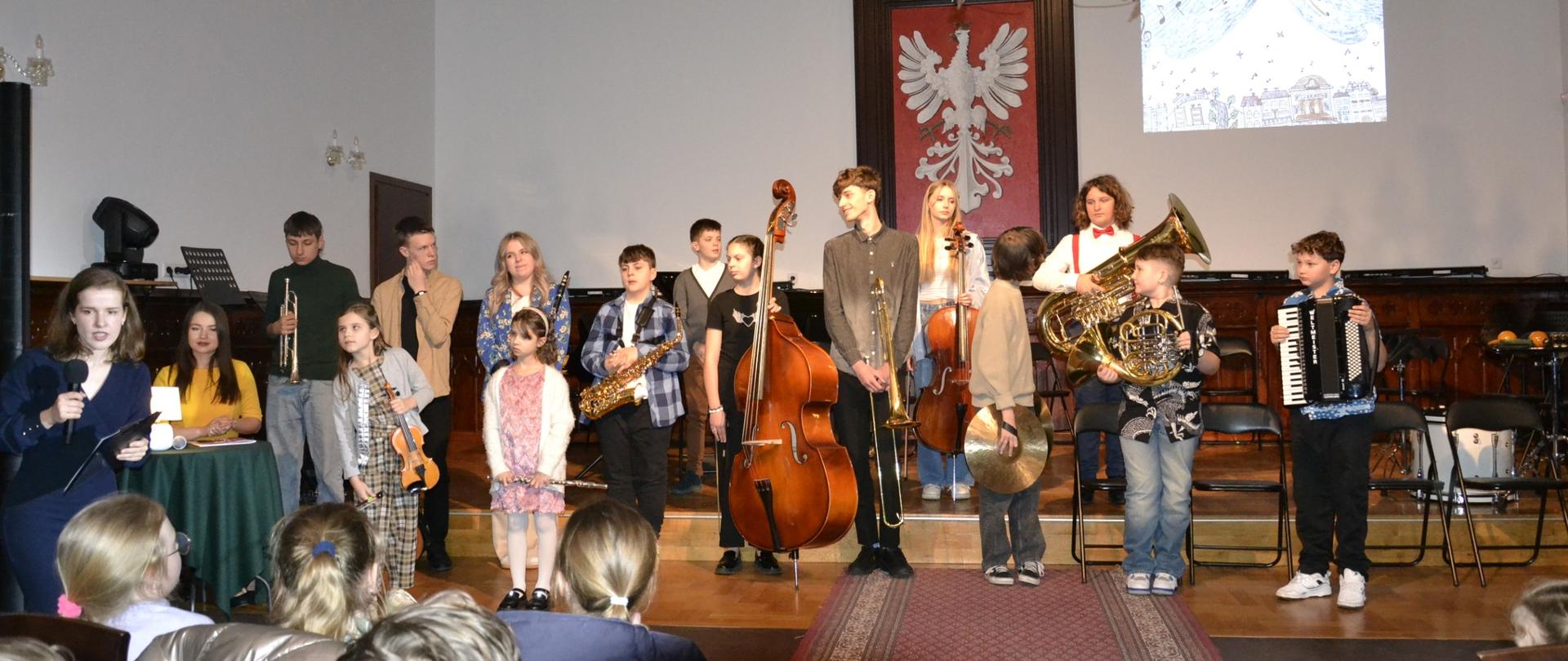 Na zdjęciu widzimy grupę młodych muzyków ze Szkoły Muzycznej w Mielcu podczas koncertu w Sali Królewskiej, który odbył się 18 marca 2025 roku. Uczniowie prezentują różnorodne instrumenty, takie jak kontrabas, saksofon, waltornia, akordeon, puzon, trąbka, wiolonczela oraz talerze perkusyjne.
