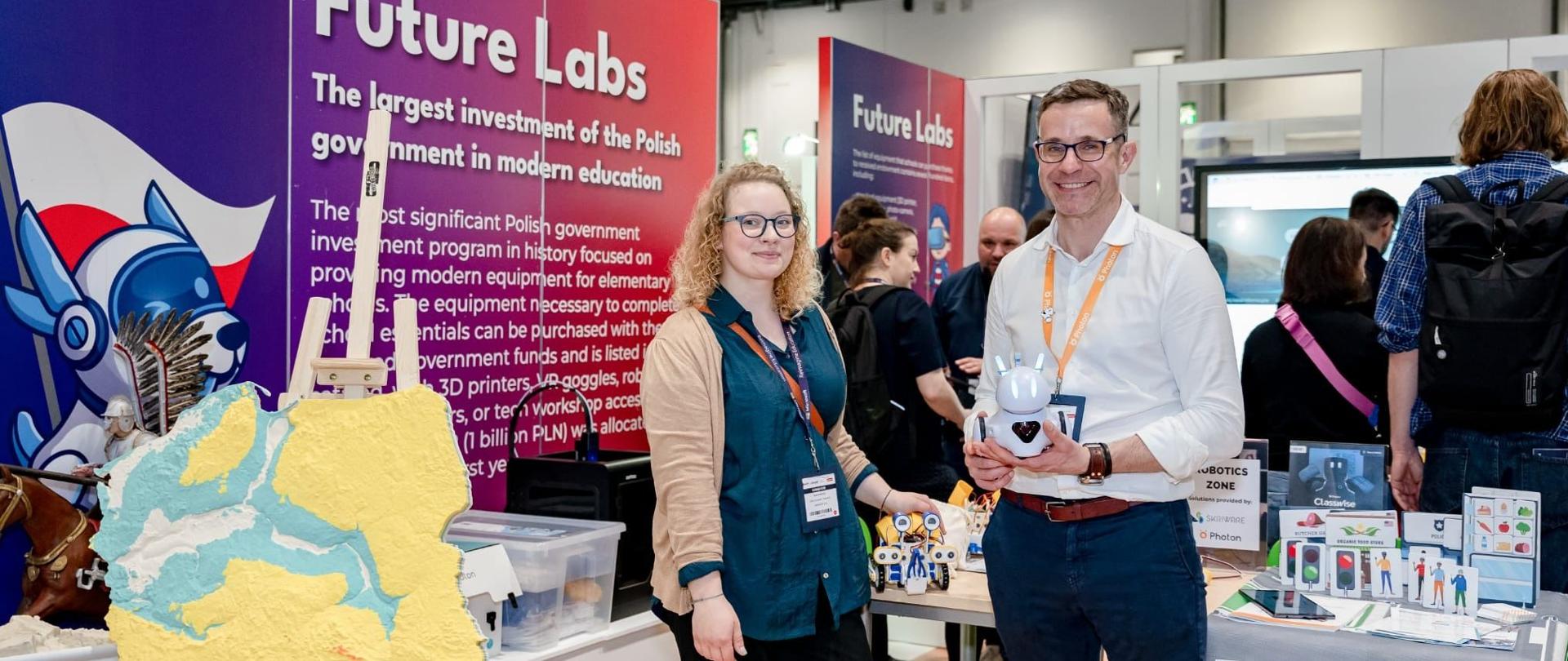 Widok na stoisko z fioletowo-różową ścianką z napisem Future Labs, przed nią stoi kobieta w niebieskiej bluzce i mężczyzna w białej koszuli, obok nich na sztalugach plastyczna mapa Polski.