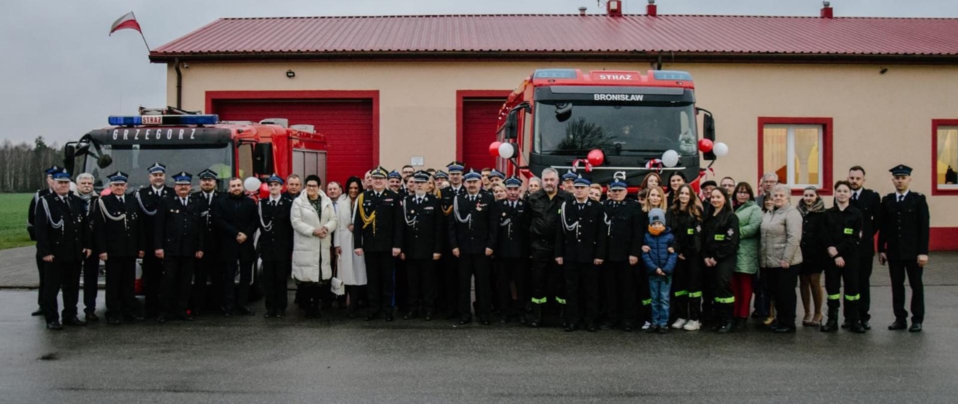 uczestnicy zebrania, druhowie strażacy przedstawiciele władzy samorządowej pozują do zdjęcia na tle samochodów i remizy osp