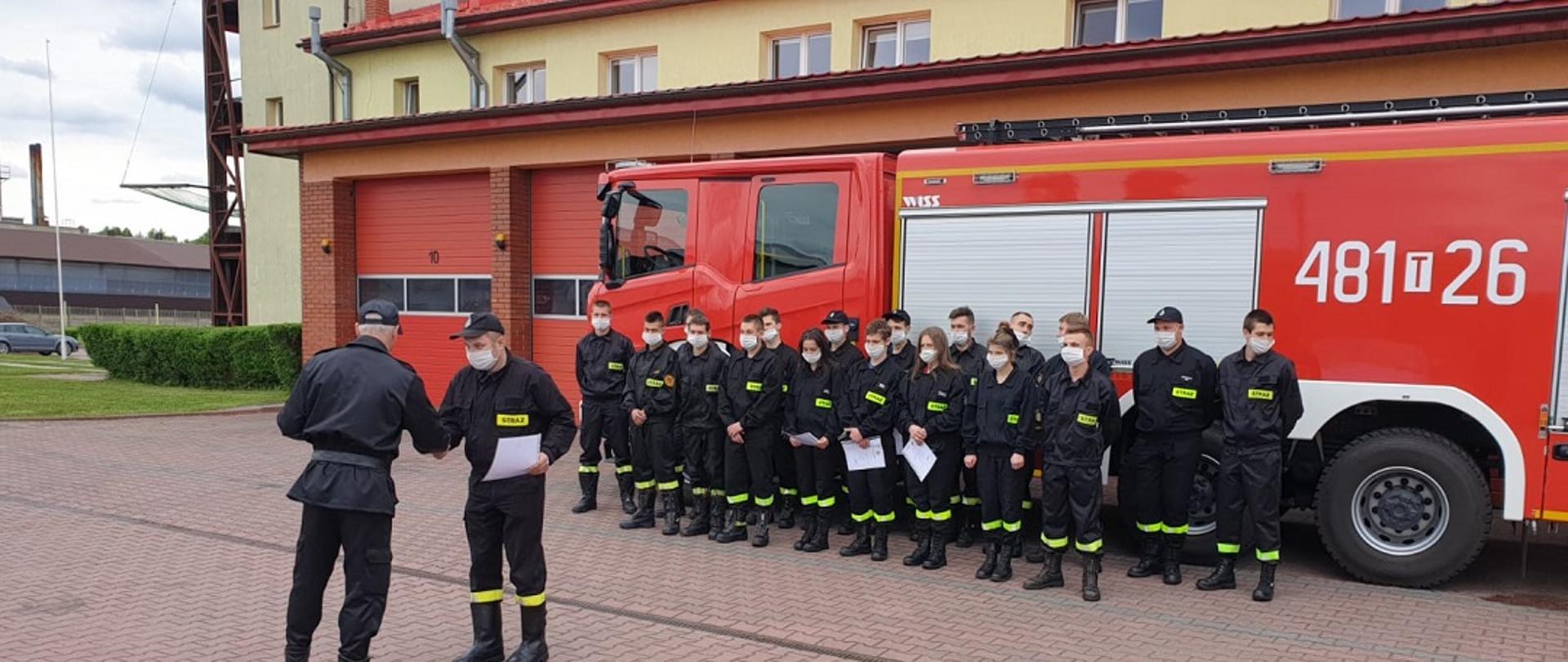 Na zdjęciu widoczna jest grupa strażaków ubranych w ubrania koszarowe koloru czarnego. Grupa strażaków stoi na tle czerwonego strażackiego samochodu gaśniczego. Na pierwszym planie na zdjęciu widoczny jest strażak ubrany w czarne ubranie dowódczo sztabowe odwrócony tyłem. Naprzeciwko niego stoi inny strażak również ubrany w czarne ubranie koszarowe. Strażacy podają sobie ręce. W tle widać jest część budynku strażnicy w kolorach czerwony i żółtym. Na dalszym planie widoczne są fragmenty budynków mieszkalnych i drzewa.