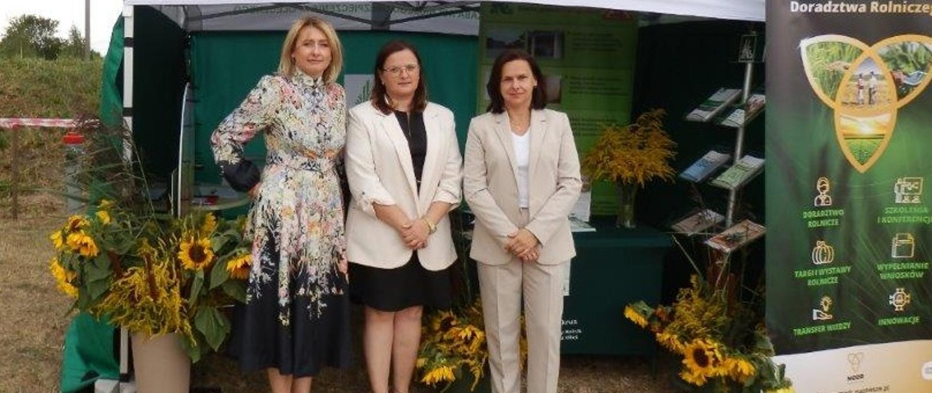 Trzy kobiety stoją przed zielonym namiotem KRUS, w którym zorganizowano stoisko informacyjno- prewencyjne dla uczestników dożynek. W namiocie z lewej strony ustawiony jest stojak z ulotkami. Namiot ozdobiono kwiatami.