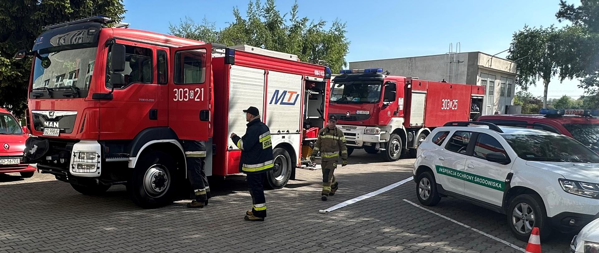 Strażacy podczas ćwiczeń: dwa wozy ratownicze PSP na parkingu siedziby WIOŚ w Gdańsku. Trzech strażaków przy wozach, na ziemi rozłożony wąż strażacki. Po lewej stronie oznakowany pojazd służbowy Inspekcji Ochrony Środowiska
