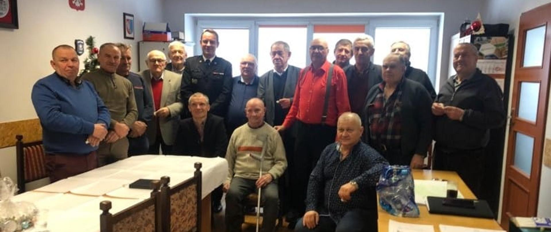 Zdjęcie wykonane w jednym z pomieszczeń jednostki ratowniczo - gaśniczej Państwowej Straży Pożarnej numer 3 w Radomiu. Na zdjęciu widać członków powiatowego koła związku emerytów i rencistów pożarnictwa rp. Mężczyźni pozują do wspólnego pamiątkowego zdjęcia. Pomiędzy nimi w drugim rzędzie stoi komendant miejski Państwowej Straży Pożarnej w Radomiu starszy brygadier Bartosz Musiał. Funkcjonariusz ubrany jest w mundur galowy w ciemnym kolorze. 